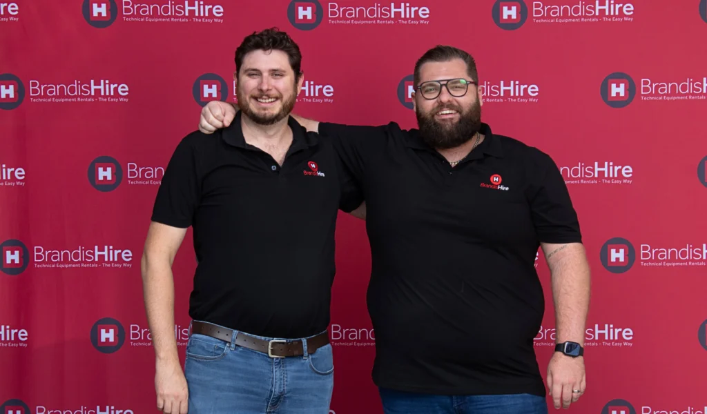 Two men in black BrandisHire polos pose with arms around each other in front of a red BrandisHire backdrop.