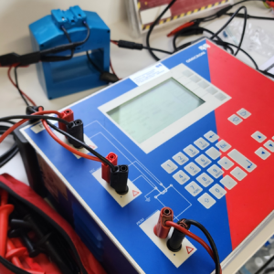 Close-up of a blue Omicron testing instrument with a blank LCD screen and red/black probe clips connected to terminals at the front panel.