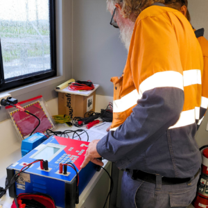 Two technicians in orange safety vests testing electrical equipment at a workbench with cables and a battery charger.